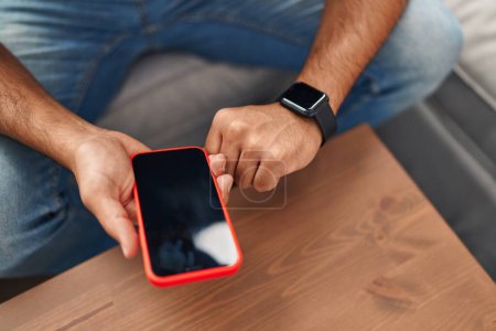 Photo for Young hispanic man using smartphone and smartwatch at home - Royalty Free Image