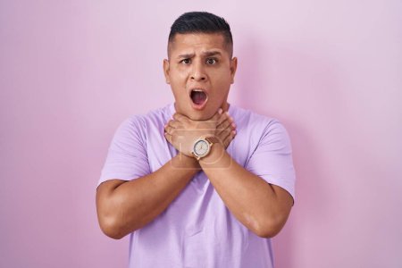 Foto de Un joven hispano parado sobre un fondo rosado gritando sofocado por un estrangulamiento doloroso. problema de salud. concepto de asfixia y suicidio. - Imagen libre de derechos