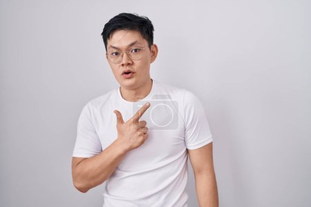 Téléchargez les photos : Young asian man standing over white background surprised pointing with finger to the side, open mouth amazed expression. - en image libre de droit