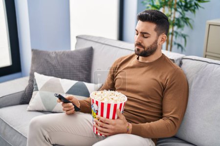 Téléchargez les photos : Jeune homme hispanique ennuyeux regarder un film assis sur le canapé à la maison - en image libre de droit