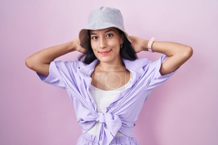 Foto de Mujer hispana joven de pie sobre fondo rosa con sombrero relajante y estiramiento, brazos y manos detrás de la cabeza y el cuello sonriendo feliz - Imagen libre de derechos