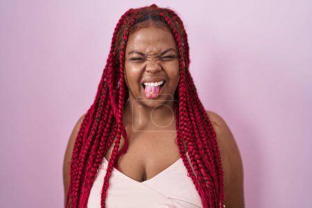 Téléchargez les photos : Femme afro-américaine avec des cheveux tressés debout sur fond rose langue collante heureux avec une expression drôle. concept d'émotion. - en image libre de droit