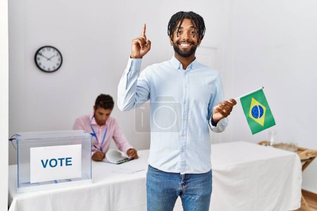 Foto de Young hispanic men at political campaign election holding brazil flag surprised with an idea or question pointing finger with happy face, number one - Imagen libre de derechos