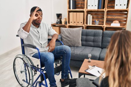 Foto de Hombre afroamericano haciendo terapia sentado en silla de ruedas haciendo buen gesto conmocionado con la cara sorprendida, los ojos mirando a través de los dedos. expresión incrédula. - Imagen libre de derechos