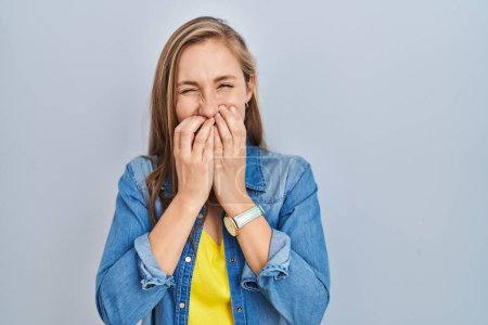 Téléchargez les photos : Jeune femme blonde debout sur fond bleu riant et embarrassé rire couvrant la bouche avec les mains, commérages et concept de scandale - en image libre de droit