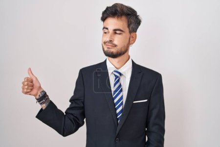 Foto de Joven hombre hispano con tatuajes vistiendo traje de negocios y corbata luciendo orgulloso, sonriendo haciendo un gesto hacia un lado - Imagen libre de derechos