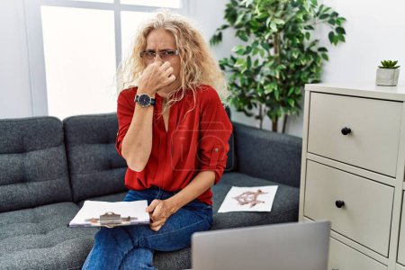Téléchargez les photos : Psychologue d'âge moyen femme au bureau de consultation sentant quelque chose puant et dégoûtant, odeur intolérable, retenant souffle avec les doigts sur le nez. mauvaise odeur - en image libre de droit