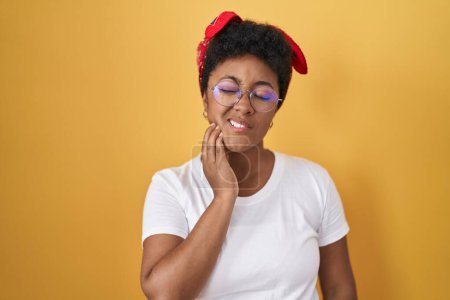 Téléchargez les photos : Jeune femme afro-américaine debout sur fond jaune touchant la bouche avec la main avec une expression douloureuse à cause de maux de dents ou de maladies dentaires sur les dents. dentiste - en image libre de droit