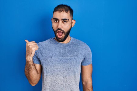 Foto de Hombre de Oriente Medio con barba de pie sobre fondo azul sorprendido señalando con el dedo de la mano a un lado, boca abierta expresión asombrada. - Imagen libre de derechos