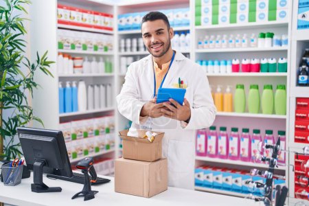 Photo for Young hispanic man pharmacist using touchpad holding pills bottle at pharmacy - Royalty Free Image