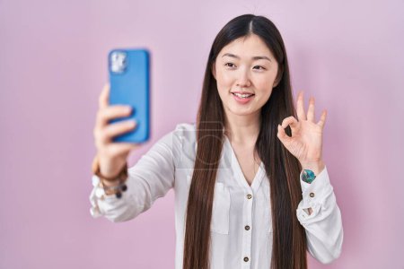 Téléchargez les photos : Chinois jeune femme prenant une photo selfie avec smartphone faire ok signe avec les doigts, sourire geste amical excellent symbole - en image libre de droit