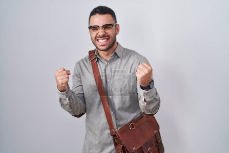 Téléchargez les photos : Jeune homme hispanique portant une valise très heureux et excité faisant geste gagnant avec les bras levés, souriant et criant pour le succès. concept de célébration. - en image libre de droit