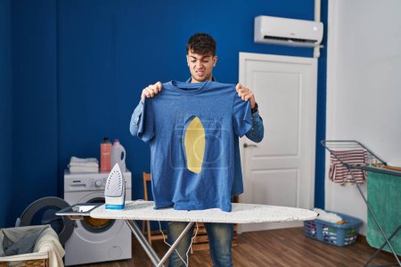 Foto de Joven hombre hispano planchando con camisa de hierro quemada en la lavandería expresión despistada y confusa. concepto de duda. - Imagen libre de derechos