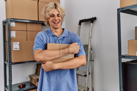 Téléchargez les photos : Jeune homme blond commerce électronique travailleur d'affaires étreignant paquet au bureau - en image libre de droit