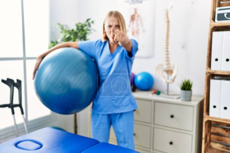 Photo for Young caucasian physiotherapist woman holding pilates ball pointing with finger to the camera and to you, confident gesture looking serious - Royalty Free Image