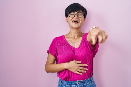 Foto de Young asian woman with short hair standing over pink background laughing at you, pointing finger to the camera with hand over body, shame expression - Imagen libre de derechos