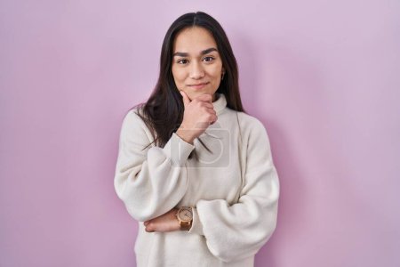 Photo for Young south asian woman standing over pink background looking confident at the camera smiling with crossed arms and hand raised on chin. thinking positive. - Royalty Free Image