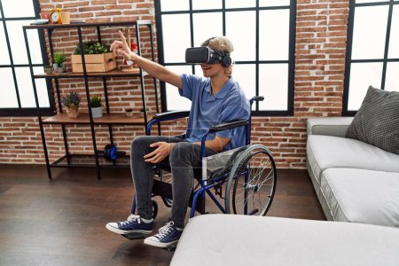 Téléchargez les photos : Jeune homme blond jouant à un jeu vidéo en utilisant des lunettes de réalité virtuelle assis sur un fauteuil roulant à la maison - en image libre de droit