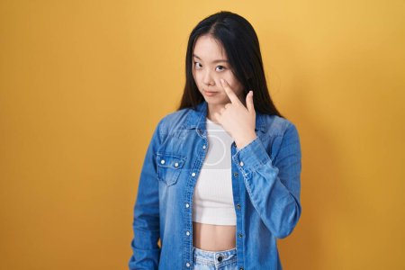Téléchargez les photos : Jeune femme asiatique debout sur fond jaune pointant vers l'œil vous regardant geste, expression suspecte - en image libre de droit