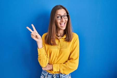 Foto de Young woman wearing glasses standing over blue background smiling with happy face winking at the camera doing victory sign. number two. - Imagen libre de derechos