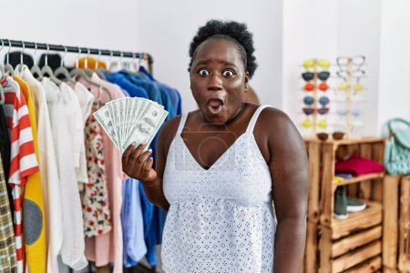 Foto de Mujer africana joven sosteniendo billetes de dólares en la tienda minorista asustada y sorprendida con la boca abierta para sorpresa, cara de incredulidad - Imagen libre de derechos