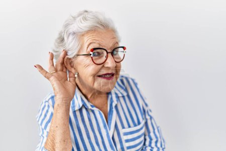 Téléchargez les photos : Femme âgée avec les cheveux gris debout sur fond blanc souriant avec la main sur l'oreille en écoutant une rumeur ou des commérages. concept de surdité. - en image libre de droit