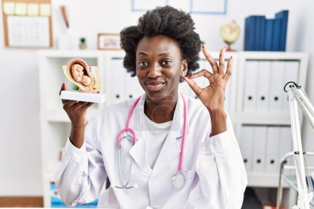 Photo for African doctor woman holding anatomical model of female uterus with fetus doing ok sign with fingers, smiling friendly gesturing excellent symbol - Royalty Free Image