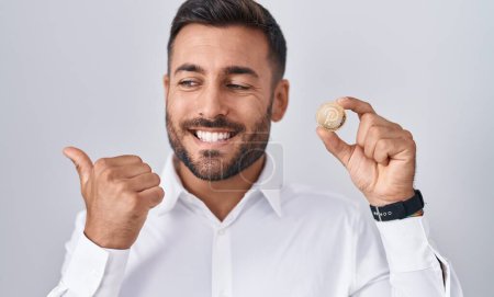Foto de Hombre hispano guapo sosteniendo moneda criptomoneda polkadot apuntando pulgar hacia el lado sonriendo feliz con la boca abierta - Imagen libre de derechos