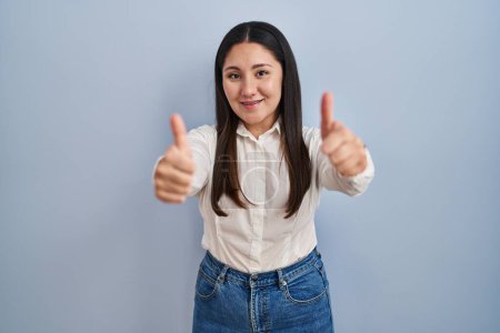 Téléchargez les photos : Young latin woman standing over blue background approving doing positive gesture with hand, thumbs up smiling and happy for success. winner gesture. - en image libre de droit