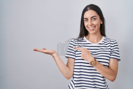 Foto de Joven morena vistiendo camiseta a rayas asombrada y sonriendo a la cámara mientras se presenta con la mano y señalando con el dedo. - Imagen libre de derechos