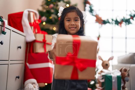Foto de Adorable chica hispana sosteniendo regalo de pie junto al árbol de Navidad en casa - Imagen libre de derechos