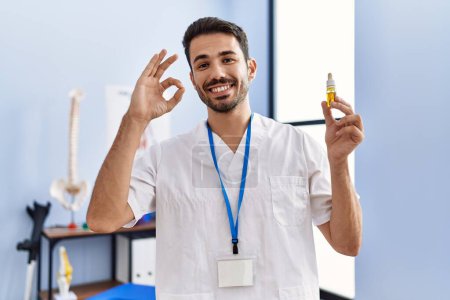 Photo for Young hispanic physiotherapist man holding cbd oil as pain relief doing ok sign with fingers, smiling friendly gesturing excellent symbol - Royalty Free Image