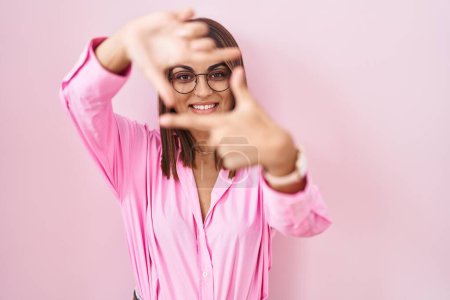Téléchargez les photos : Jeune femme hispanique portant des lunettes debout sur fond rose souriant faisant cadre avec les mains et les doigts avec le visage heureux. concept de créativité et photographie. - en image libre de droit