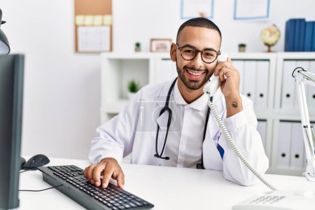 Photo for Young hispanic man wearing doctor uniform speaking on the phone at clinic - Royalty Free Image