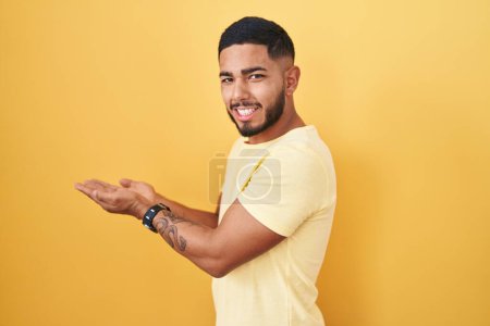 Foto de Joven hombre hispano de pie sobre fondo amarillo señalando a un lado con las manos abiertas palmeras mostrando espacio de copia, presentando anuncio sonriente emocionado feliz - Imagen libre de derechos