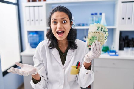 Photo for Hispanic woman with dark hair working at scientist laboratory holding money celebrating victory with happy smile and winner expression with raised hands - Royalty Free Image