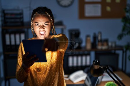 Foto de Mujer afroamericana con trenzas trabajando en la oficina por la noche con la tableta señalando disgustado y frustrado a la cámara, enojado y furioso con usted - Imagen libre de derechos