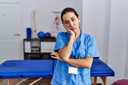 Foto de Mujer hispana joven con uniforme de fisioterapeuta de pie en la clínica pensando que se ve cansada y aburrida con problemas de depresión con los brazos cruzados. - Imagen libre de derechos