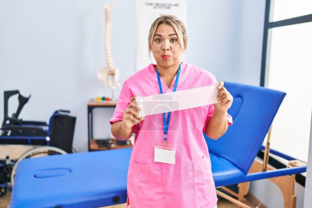 Foto de Young hispanic woman working at pain recovery clinic holding bandage puffing cheeks with funny face. mouth inflated with air, catching air. - Imagen libre de derechos