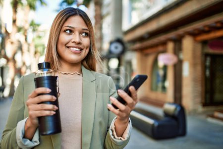 Téléchargez les photos : Jeune femme latine souriant confiant en utilisant smartphone à la rue - en image libre de droit