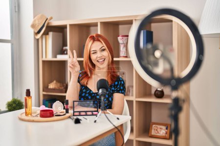 Téléchargez les photos : Jeune femme caucasienne appliquant maquillage faire tutoriel souriant avec un clin d'oeil visage heureux à la caméra faisant signe de victoire. numéro deux. - en image libre de droit
