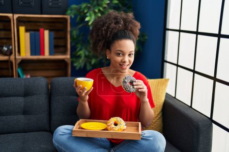Foto de Young african american woman having breakfast sitting on sofa at home - Imagen libre de derechos