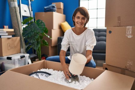 Photo for Young beautiful hispanic woman sitting on floor unboxing cardboard box at new home - Royalty Free Image