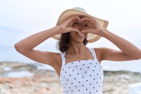 Foto de Young african american woman wearing summer hat doing heart gesture with hands at seaside - Imagen libre de derechos