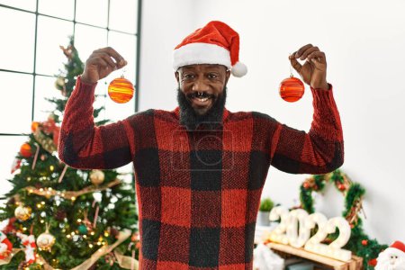 Foto de Joven afroamericano hombre sonriendo confiado sosteniendo bolas de pie por sombrero de Navidad en casa - Imagen libre de derechos