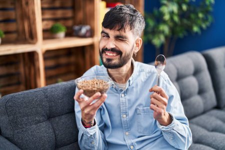 Foto de Joven hombre hispano con barba comiendo cereales integrales sanos guiñando el ojo mirando a la cámara con expresión sexy, cara alegre y feliz. - Imagen libre de derechos