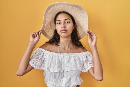 Téléchargez les photos : Young brazilian woman wearing summer hat over yellow background relaxed with serious expression on face. simple and natural looking at the camera. - en image libre de droit