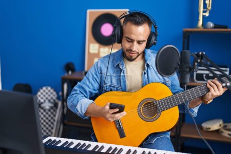 Foto de Joven artista hispano usando smartphone tocando guitarra clásica en estudio de música - Imagen libre de derechos