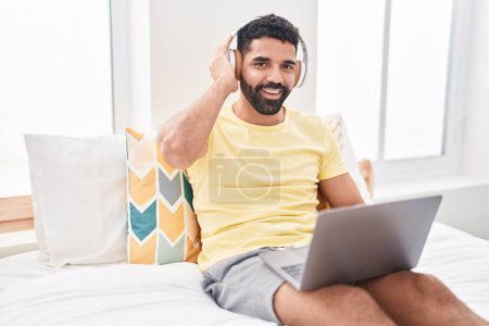 Téléchargez les photos : Young arab man using laptop and headphones sitting on bed at bedroom - en image libre de droit
