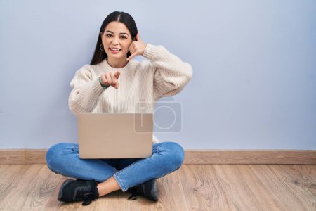 Foto de Young woman using laptop sitting on the floor at home smiling doing talking on the telephone gesture and pointing to you. call me. - Imagen libre de derechos